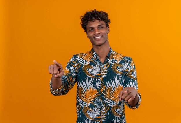 Sonriente joven guapo de piel oscura con cabello rizado en hojas camisa estampada apuntando a la cámara con los dedos índices sobre un fondo naranja