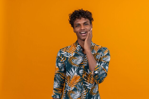 Sonriente joven guapo de piel oscura con cabello rizado en hojas camisa estampada sosteniendo la mano en la mejilla sobre un fondo naranja