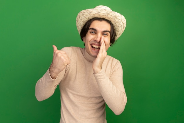 Sonriente joven guapo con puntos de sombrero al lado poniendo la mano alrededor de la boca aislada en la pared verde con espacio de copia