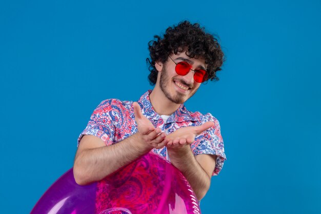 Sonriente joven guapo rizado con gafas de sol sosteniendo el anillo de natación estirando las manos sobre la pared azul aislada con espacio de copia