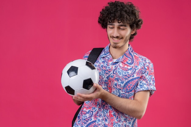 Sonriente joven guapo rizado sosteniendo un balón de fútbol y mirándolo en la pared rosa aislada con espacio de copia