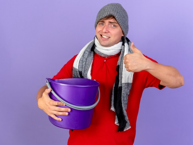 Sonriente joven guapo rubio enfermo con gorro de invierno y bufanda sosteniendo un cubo de plástico mirando a la cámara mostrando el pulgar hacia arriba aislado sobre fondo púrpura