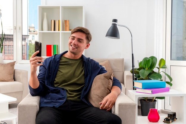 Sonriente joven guapo rubio se sienta en un sillón sosteniendo y mirando el teléfono