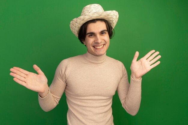 Sonriente joven guapo con sombrero extendiendo las manos aisladas en la pared verde