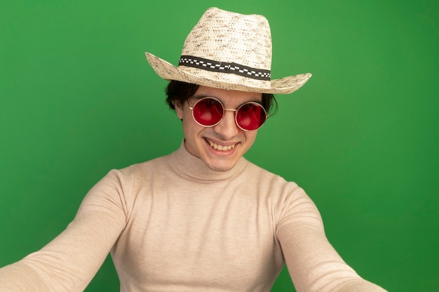 Foto gratuita sonriente joven guapo con sombrero con gafas aislado en la pared verde