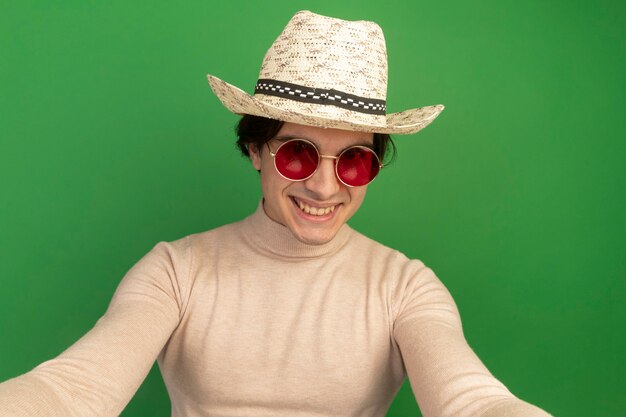 Sonriente joven guapo con sombrero con gafas aislado en la pared verde