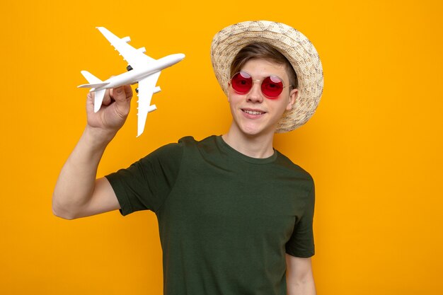 Sonriente joven guapo con sombrero con gafas sosteniendo avión de juguete aislado en la pared naranja