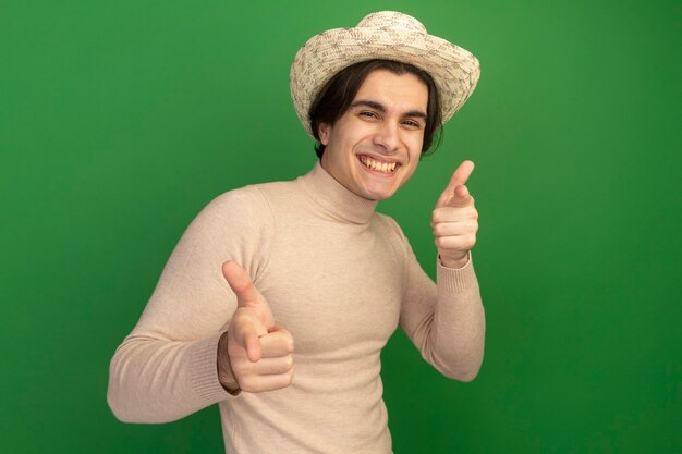 Sonriente joven guapo con sombrero que le muestra gesto aislado en la pared verde