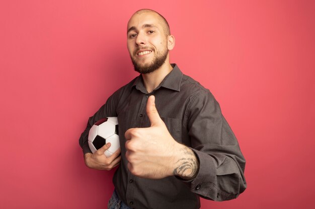 Sonriente joven guapo sosteniendo la bola y mostrando el pulgar hacia arriba