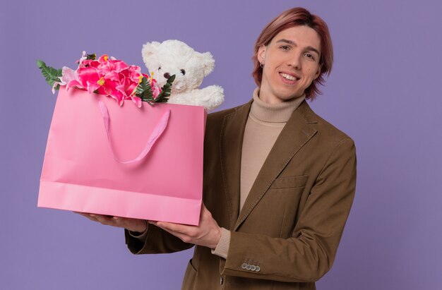 Sonriente joven guapo sosteniendo una bolsa de regalo rosa con flores y osito de peluche