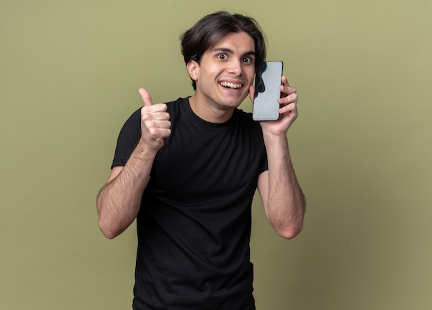 Sonriente joven guapo vistiendo camiseta negra con reloj despertador alrededor de la cara mostrando el pulgar hacia arriba aislado en la pared verde oliva