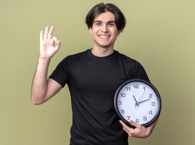 Sonriente joven guapo vistiendo camiseta negra con reloj de pared mostrando gesto bien aislado en la pared verde oliva