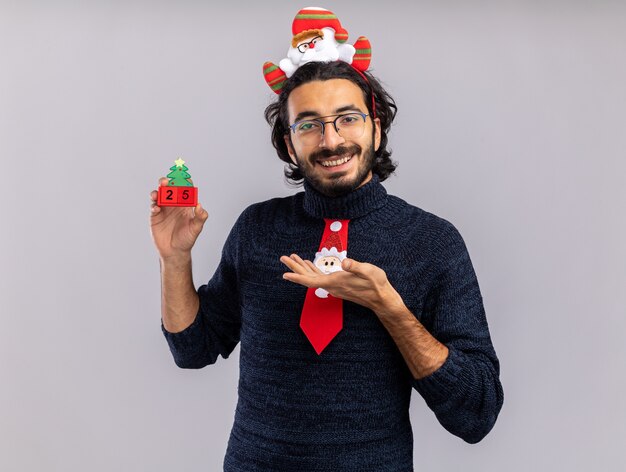 Sonriente joven guapo vistiendo corbata de navidad con aro de pelo sosteniendo y puntos en juguete de navidad aislado sobre fondo blanco.