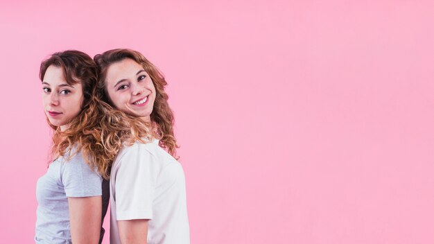 Sonriente joven hermana de pie espalda con espalda sobre fondo rosa