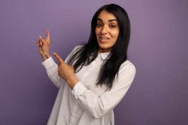 Sonriente joven hermosa chica con camiseta blanca apunta detrás aislado en púrpura con espacio de copia