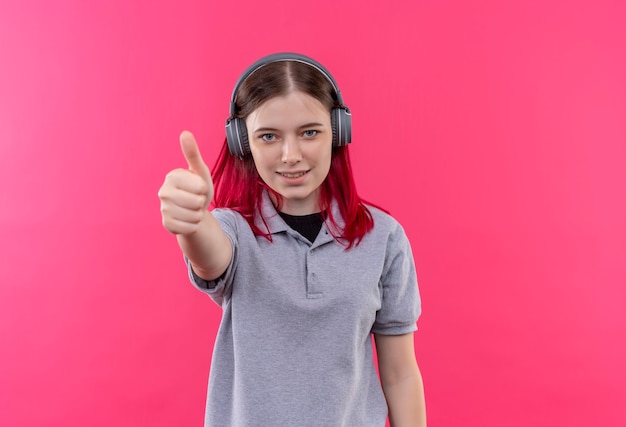 Sonriente joven hermosa chica con camiseta gris en auriculares con el pulgar hacia arriba sobre fondo rosa aislado con espacio de copia