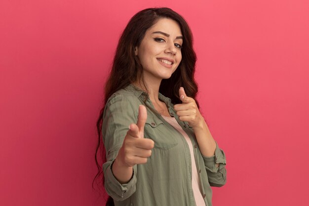 Sonriente joven hermosa chica con camiseta verde oliva puntos en la cámara aislada en la pared rosa