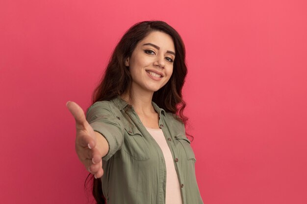 Sonriente joven hermosa chica con camiseta verde oliva tendiendo la mano a la cámara aislada en la pared rosa con espacio de copia