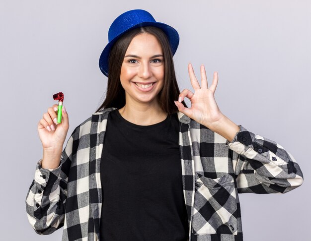 Sonriente joven hermosa chica con sombrero de fiesta sosteniendo un silbato de fiesta mostrando un gesto bien aislado en la pared blanca
