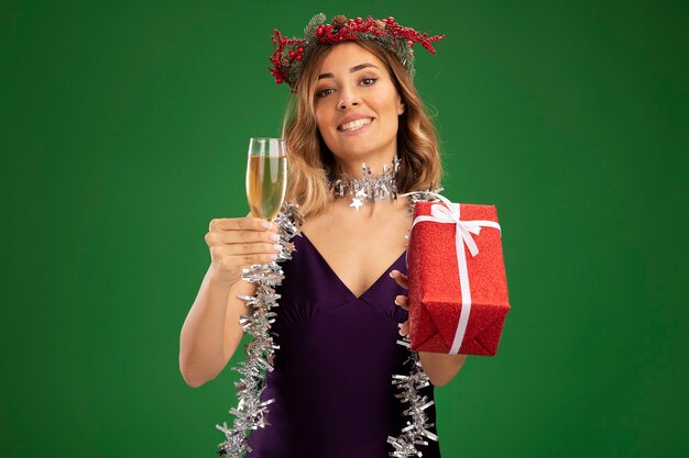 Sonriente joven hermosa chica con vestido púrpura y guirnalda wirh garland en el cuello sosteniendo una copa de champán con caja de regalo aislada sobre fondo verde