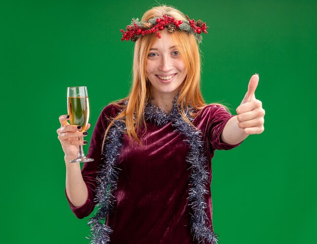 Sonriente a joven hermosa chica con vestido rojo con corona y guirnalda en el cuello sosteniendo una copa de champán mostrando el pulgar hacia arriba aislado sobre fondo verde