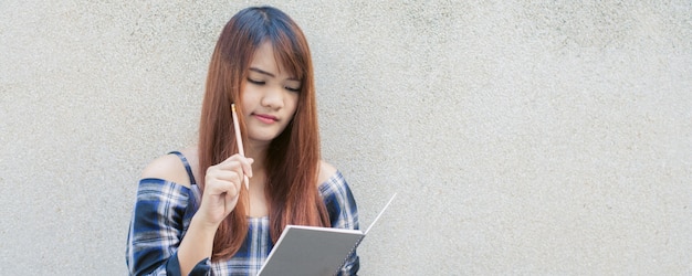 Sonriente joven hermosa mujer asiática pensando con libro de escritura sobre fondo de muro de hormigón. Imágenes de estilo de efecto vintage.