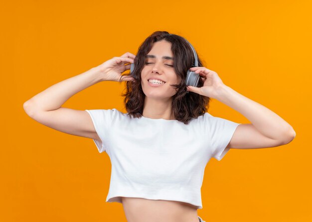 Sonriente joven hermosa mujer con auriculares y poner las manos sobre ellos con los ojos cerrados en la pared naranja aislada