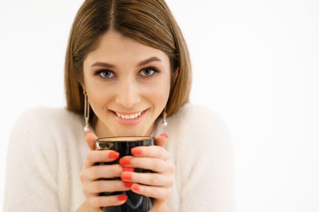 Sonriente joven hermosa mujer feliz con cabello largo disfrutando de capuchino sobre fondo blanco. Mujer de belleza disfrutando de café. Taza de bebida caliente.