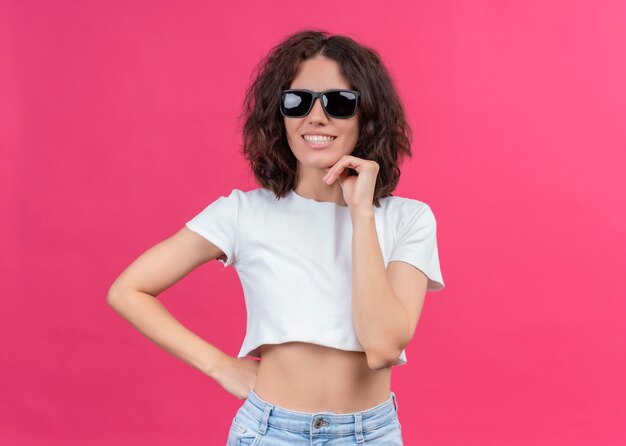 Sonriente joven hermosa mujer con gafas de sol y poniendo la mano en la barbilla en la pared rosa aislada con espacio de copia