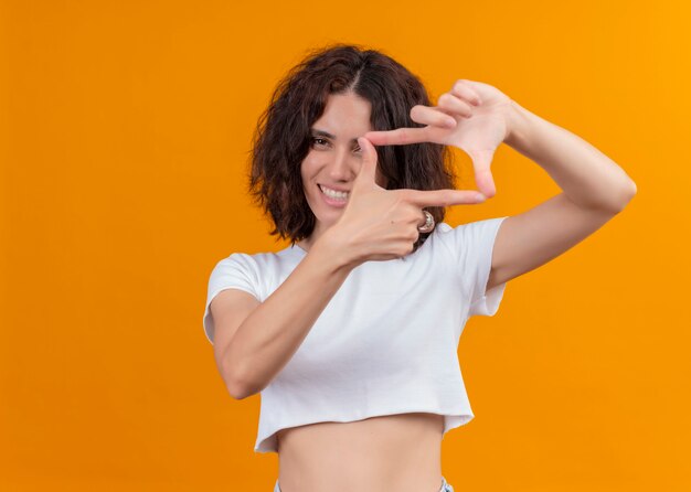 Sonriente joven hermosa mujer haciendo gesto de marco en la pared naranja aislada con espacio de copia