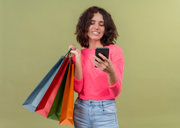 Sonriente joven hermosa mujer sosteniendo bolsas de cartón y teléfono móvil en la pared verde aislada con espacio de copia