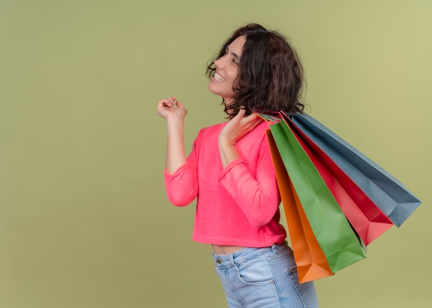 Sonriente joven hermosa mujer sosteniendo bolsas de papel de pie en la vista de perfil y mirando al lado izquierdo en la pared verde aislada con espacio de copia