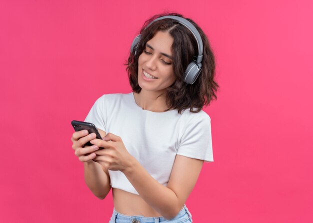 Sonriente joven hermosa mujer usando audífonos y sosteniendo el teléfono móvil en la pared rosa con espacio de copia