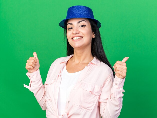 Sonriente joven hermosa mujer vistiendo gorro de fiesta mostrando los pulgares para arriba