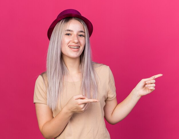 Sonriente joven hermosa mujer vistiendo gorro de fiesta con puntos de aparatos dentales en el lado aislado en la pared rosa