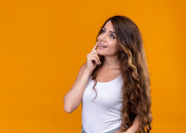 Sonriente joven hermosa niña mirando hacia el lado izquierdo y poniendo el dedo en la mejilla en el espacio naranja aislado con espacio de copia