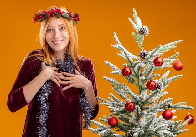 Sonriente joven hermosa niña de pie cerca del árbol de navidad con vestido rojo y corona con guirnalda en el cuello poniendo las manos sobre sí misma aislada sobre fondo naranja