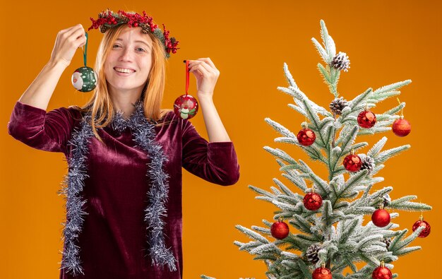 Sonriente joven hermosa niña de pie cerca del árbol de Navidad con vestido rojo y corona con guirnalda en el cuello sosteniendo bolas de Navidad aisladas sobre fondo naranja