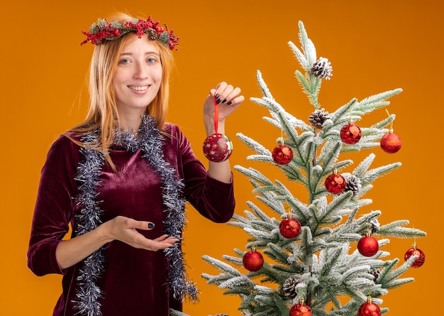 Sonriente joven hermosa niña de pie cerca del árbol de Navidad con vestido rojo y corona con guirnalda en el cuello sosteniendo bolas de Navidad aisladas sobre fondo naranja