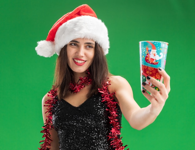 Sonriente joven hermosa niña con sombrero de navidad con guirnalda en el cuello sosteniendo la taza de navidad aislado en la pared verde