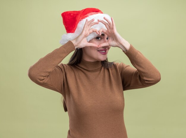 Sonriente joven hermosa niña con sombrero de navidad mostrando gesto de corazón aislado sobre fondo verde oliva