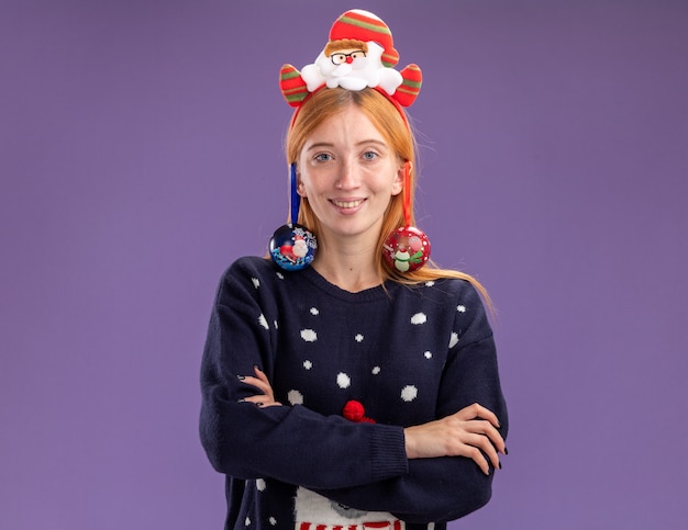 Sonriente joven hermosa niña con suéter de Navidad con aro de pelo de Navidad colgado bolas de Navidad en la oreja cruzando las manos aisladas sobre fondo púrpura