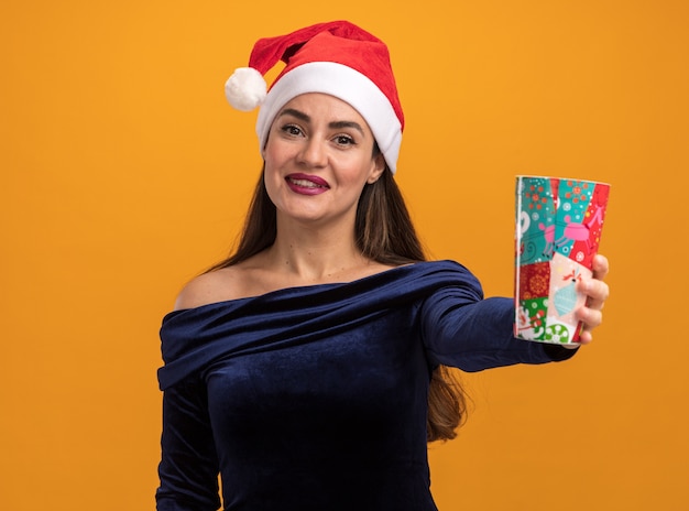 Sonriente joven hermosa niña con vestido azul y sombrero de navidad sosteniendo la taza de navidad aislado en la pared naranja