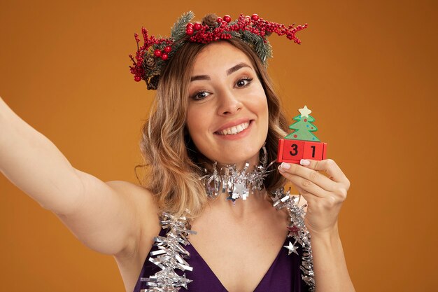 Sonriente joven hermosa niña con vestido púrpura y corona con guirnalda en el cuello sosteniendo juguetes de Navidad y cámara aislada sobre fondo marrón