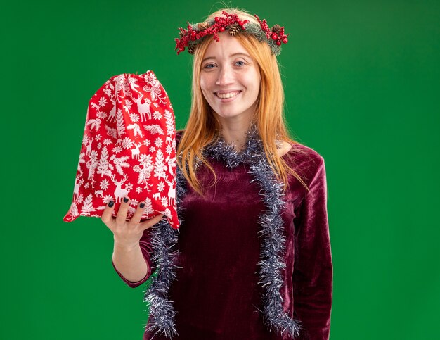 Sonriente joven hermosa niña con vestido rojo con corona y guirnalda en el cuello sosteniendo una bolsa de Navidad aislada sobre fondo verde