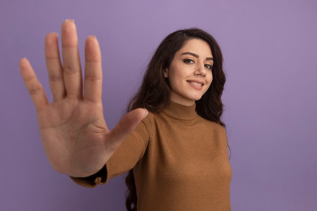 Foto gratuita sonriente joven hermosa niña vistiendo un suéter de cuello alto marrón mostrando cinco aislados en la pared púrpura