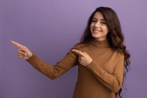 Sonriente joven hermosa niña vistiendo suéter de cuello alto marrón puntos al lado aislado en la pared púrpura con espacio de copia