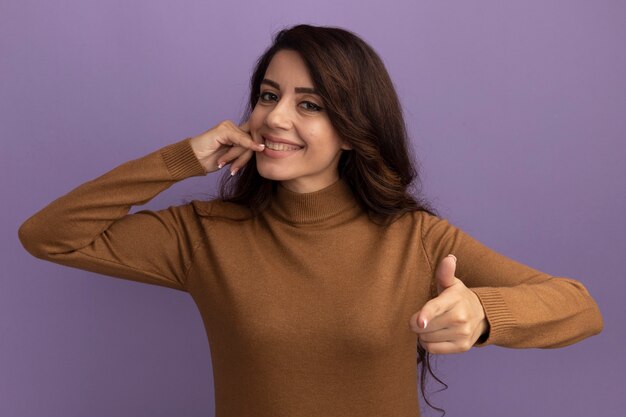 Sonriente joven hermosa niña vistiendo un suéter de cuello alto marrón que muestra el gesto de llamada telefónica y puntos aislados en la pared púrpura