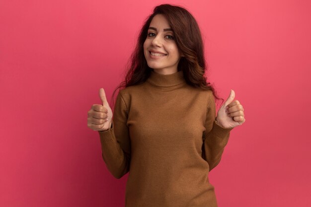 Sonriente joven hermosa niña vistiendo suéter de cuello alto mostrando los pulgares para arriba aislados en la pared rosa