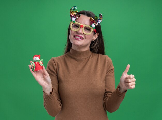 Sonriente joven hermosa niña vistiendo un suéter marrón y gafas de navidad sosteniendo un juguete de navidad mostrando el pulgar hacia arriba aislado en la pared verde